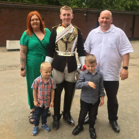 Family photos Jack stands in uniform with his family. He smiles at the camera in gold and black with his mum beside him in a green dress and dad in white. Two younger boys also stand in front of him