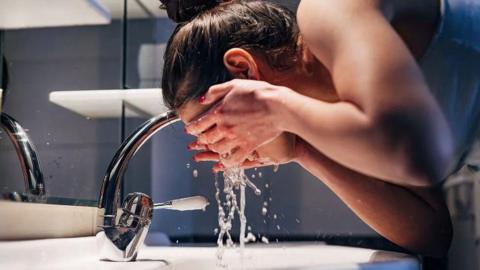 A woman stood over a sink splashing her face with water