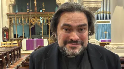 Robby West/BBC James Stewart, standing inside his church. He wears the black shirt and jacket and white dog collar. There are wooden pews behind him