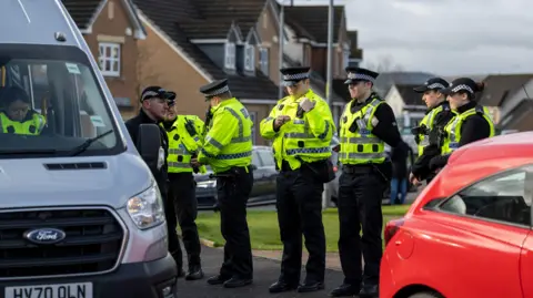 PA Media Seven police officers in high viz vests in a residential area, standing in a line next to a white police vehicle with another officer sitting inside.