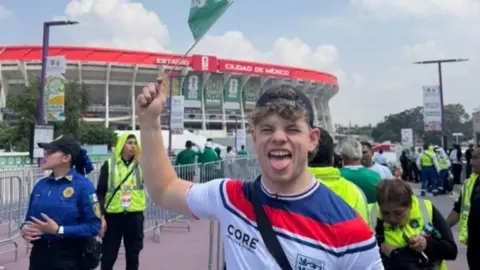A man in a white England football shirt and a black cap on backwards holds a Mexico flag and stands in front of the Mexico City Stadium. People in hi-vis stand around him in the background and a female police officer stands beside him.