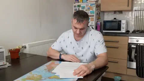 A middle-aged man looks down at some documents as he sits at a table. He is wearing a T-shirt