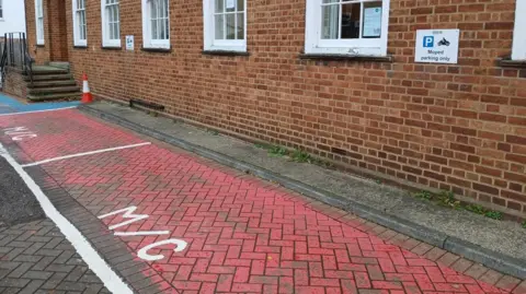 Epsom and Ewell Borough Council Two parking spaces marked with red paint with the letters M/C written in white paint. The spaces are next to a brown brick building