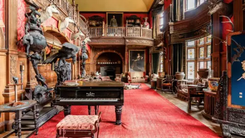 Savills The grand hall of Kinloch Castle with an air of faded grandeur with red carpet and a grand piano and numerous statues and stags heads