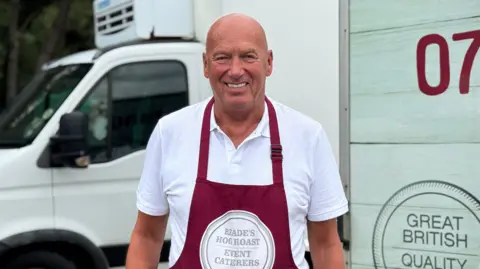 George King/BBC Kevin Reade, wearing a white polo shirt and a burgundy coloured apron, smiling into the camera.