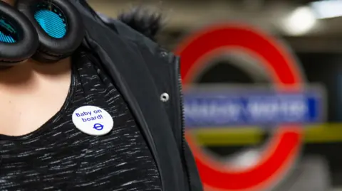 Transport for London A person wearing a "Baby on board!" badge with the Transport for London logo on their dark mottled shirt.