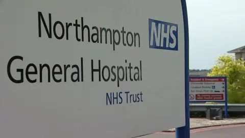 BBC Sign saying "Northampton General Hospital NHS Trust" in black and blue writing on a white background. Another sign is out of focus in the background.