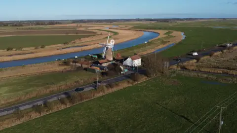 John Fairhall/BBC An aerial view of part of the A47 Acle Straight near Great Yarmouth in Norfolk. The road runs between fields, and some houses. a windpump and a river can also be seen.