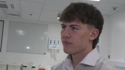 Young, second year medical student John Morris. He has dark brown hair in a short, mullet style. He is wearing a smart, stripped shirt. He is in a classroom and behind him is a blank whiteboard.