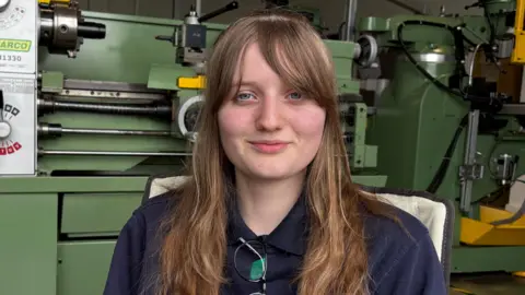 Ben Schofield/BBC Tegan Smith looking straight to camera. She is smiling slightly and is wearing a dark blue top, with a pair of glasses folded and tucked into the collar. She has light brown coloured hair, which is falling over her shoulders. Large pieces of industrial machinery can be seen in the background.