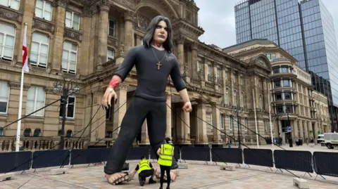 An inflatable Ozzy Osbourne outside the Birmingham Town Hall, with two workmen standing in front of him.