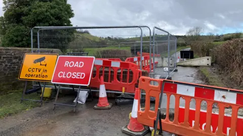 A country road surrounded by orange cones, plastic barriers and metal fencing with a stone wall visible on one side.