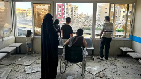 A family including a person in a wheelchair stand next to the window of the Jabal Amel hospital and look out to survey extensive damage to the neighbourhood, with broken glass and rubble on the floor of the hospital room they stand in, in Tyre on Monday.