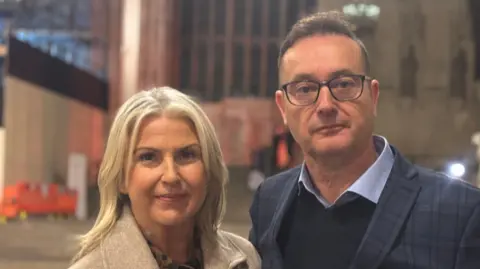 Christine Lock with blond hair wearing a cream coat (left) with husband Richard with brown hair wearing a pale blue shirt navy jumper and blue suit jacket. He has black glasses.