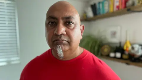 Steve Hubbard/BBC Yashpal looks directly at the camera, wearing a red T-shirt with a 