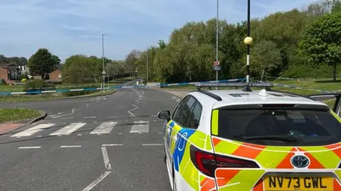 A police car can be seen in the foreground, parked before a zebra crossing with police tape stretched across it.