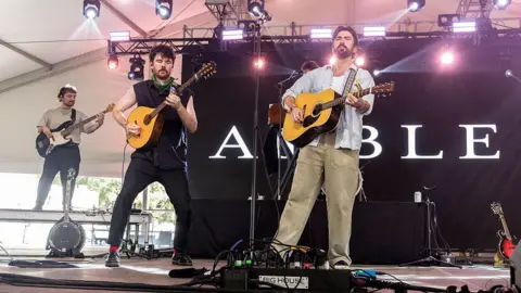 Getty Images Oison McCaffrey (L) and Robbie Cunningham of Amble perform in concert during the 2025 Austin City Limits Music Festival at Zilker Park on October 03, 2025 in Austin, Texas. All three are holding guitars and are stood in front of a large Amble backdrop.