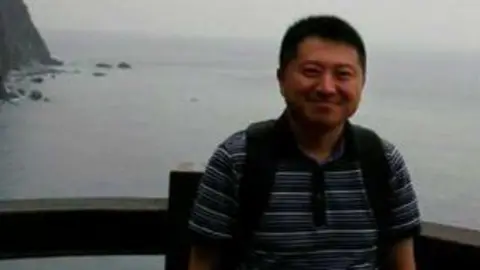Liu Hu wearing a blue polo shirt and smiling as he poses for a photo. Behind him is the sea.