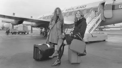 Getty Images Two woman pose in front of a plane with suitcases in a black-and-white image. One with long fair hair is sitting on her suitcase and wearing tight black boots which come over the knee and a coat with a fur-trimmed collar.l The othe rhas long brown hair and wears a trench coat, a buttoned up skirt and a black top.