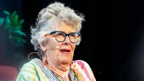 A woman with short grey hair wearing navy and orange glasses and a multicoloured stripey dress. She has a microphone on her cheek and is talking.