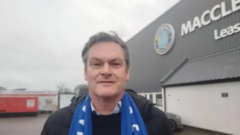 Paul Burnell/BBC Smart has short dark hair and is wearing a blue and white Macclesfield scarf and a black sweater. He is standing outside the club's stadium.