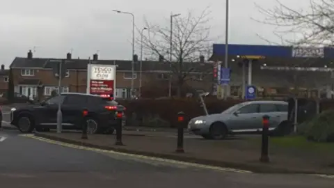 A line of traffic which involves cars exiting a Tesco Extra car park. There are two black cars and a silver car in the traffic queue. Behind the Tesco is a line of residential terraced houses. 