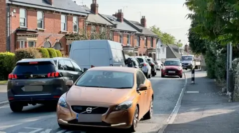 A narrow and busy road in a residential street with lots of cars passing both ways, pictured on a nice sunny day. The sunlight shines on the orange brick terraced houses that line up the road on one side, while on the other a side a pavement is clear of any pedestrians.
