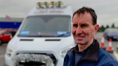 A man looks at the camera an is wearing a navy blue jacket over a blue sweater and checked shirt. He has short cark brown hair. He is stood in front of a white van decorated with silver tinsel.