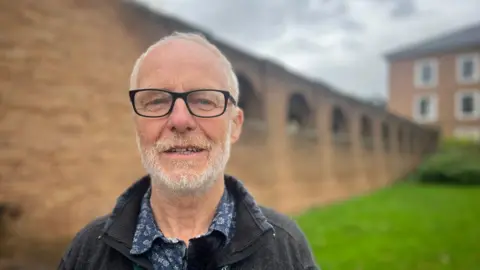 Henry Gent, wearing black-framed glasses, a blue patterned shirt and a grey jacket, with County Hall blurred in the background.