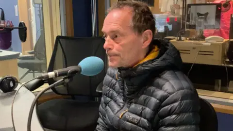 BBC A man with short brown hair, a thick black coat, sitting in a chair in a studio with a blue microphone pointing at his face.