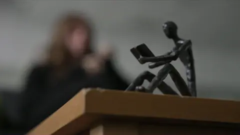 A blurred outline of a woman sitting at a coffee table. A figurine of a stick man reading a book is in the foreground.