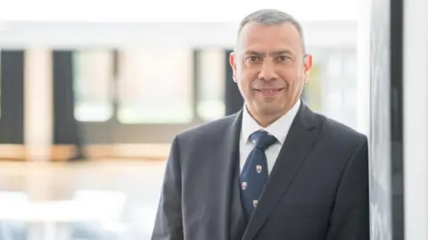 A man in a navy blazer and a white shirt and navy tie looks at the camera.     