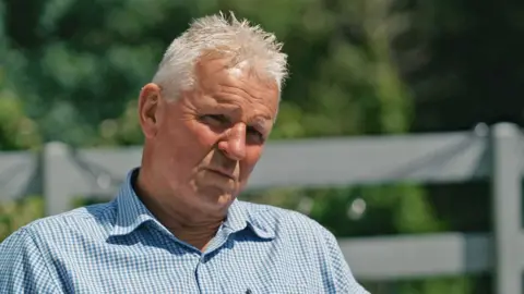 Brian Maywood, who has grey hair and a blue checked shirt, speaks to the BBC in his garden.