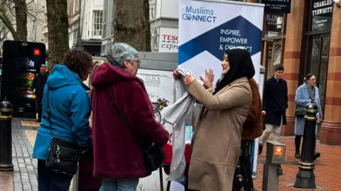 A woman wearing a black hijab and light brown long coat is talking to two ladies. One lady has curly brown hair and is wearing a blue hooded coat while the other lady has short grey hair and is wearing a red hooded fury coat.