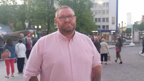 A man, with short dark hair and a tight beard, wearing a pink shirt with rolled up sleeves. There are people standing around behind him in the background.