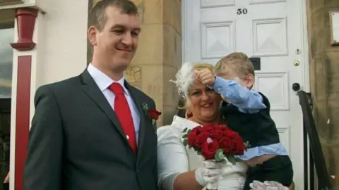 Joanne Howie is wearing a long-sleeved wedding dress with white gloves. She is holding a bouquet of red roses and a little boy. Her husband Christina Howie is standing next to her outside the white door of the registration office. He is wearing a red tie and a dark grey suit. He has short brown hair.