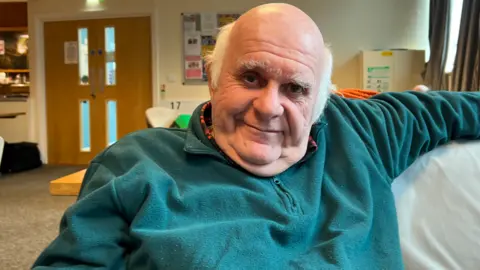 The picture shows David Greenway sitting on a sofa in Edgar Hall in Somerton. He is looking into the camera.  David is wearing a green jumper.