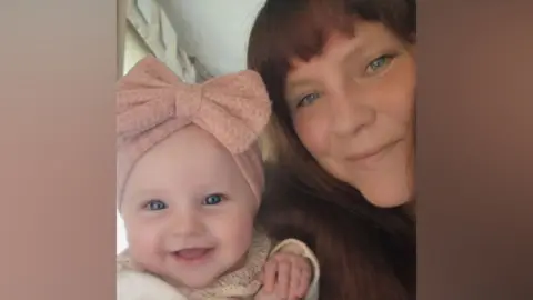 A selfie of a smiling mum with long brown hair with a fringe, and her baby daughter, who is smiling and has a large pink bow on her head.