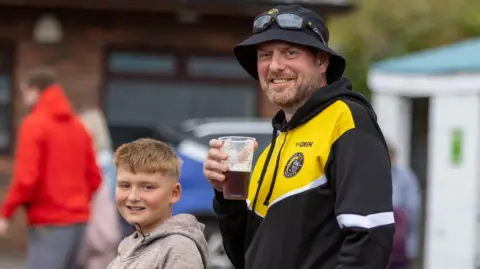 Craig Hawkhead Photography A man holding a beer and a boy stand outdoors in matching black and yellow sports hoodies at a rugby match.