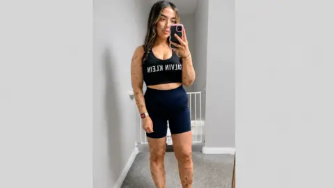 Laura Kelly Laura has long brown hair and is wearing a black top. Her arms are visible and she is taking a selfie with her phone 