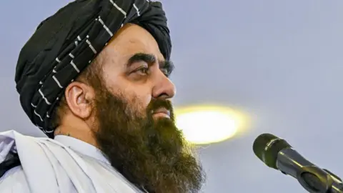 Afghanistan's Foreign Minister Amir Khan Muttaqi, with a long black beard, is in profile with a microphone directly in front of him. 