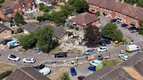 Ant Saddington/BBC Aerial view of a house which has suffered an explosion and is completely destroyed. There is nothing but rubble and debris left. There are cars and vans parked on the road in the surrounding area. There are roadworks taking place on the road near the house.