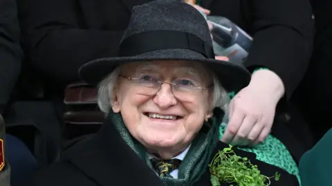 Michael D Higgins, who is wearing a dark, fedora hat, smiles at St Patricks Day celebrations kick off in Dublin. The president is an older man, with long, white hair. He is wearing a pair of rimless, rectangular glasses, a green scarf, and has a bundle of shamrock on his chest.