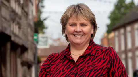 Woman smiling, wearing a red and black animal print blouse, with a blurred high street behind her