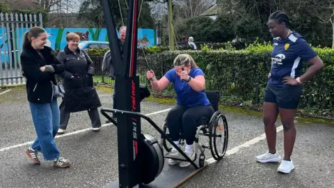 Katherine Selby Sharland in her wheelchair uses a pulling exercise machine while other people stand around her and watch on. 