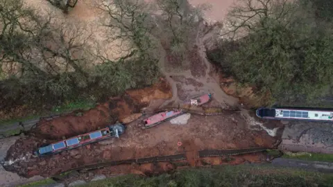PA Media An aerial view of a muddy trench with three long narrowboats in it and a large breach in the side, with brown water flowing into a nearby field