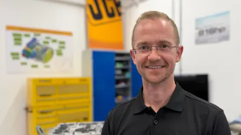 Danny Pike / BBC A man with glasses stood in a engineering workshop. A JCB sign is behind him on the wall.