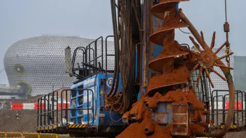 HS2 A huge drill has soil on it and stands next to large machinery with the Bullring shopping centre in the background.