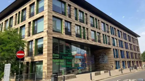 A modern council building with beige and brown stripes, colourful window frames and a large coat of arms by the entrance. The names of local towns are printed on a tall glass wall beside it, with a 'No Entry' sign street sign in the foreground.