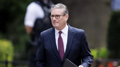 EPA/Shutterstock A man, Keir Starmer, wearing a dark coloured suit and burgundy tie. He is holding a binder in his hand, and is turning away from a lectern.